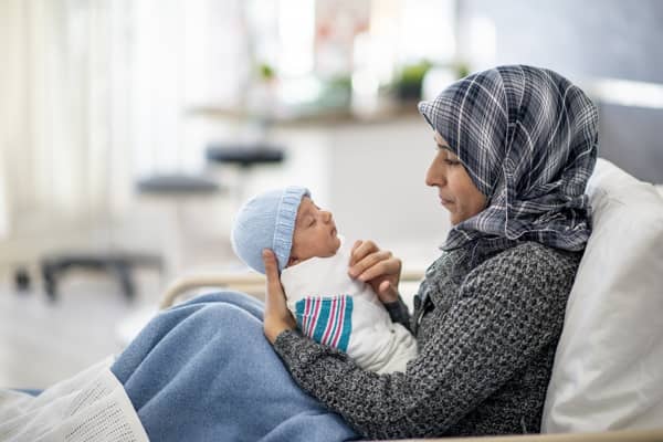 A Muslim woman is laying in a hospital bed. She is holding her newborn son in her arms. She is watching over her baby while he sleeps.